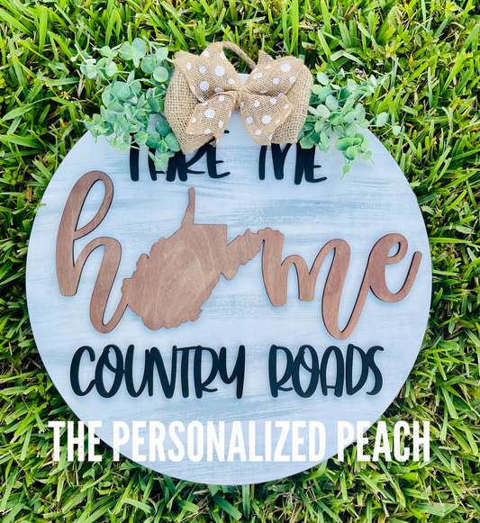 “Take me home” “country roads” with West Virginia cut through the word home. wooden door hanger with greenery and a burlap bow